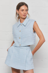 A woman is wearing a light blue sleeveless cropped vest with a classic collar and large, decorative buttons down the front. She pairs it with a matching high-waisted skirt that has a textured fabric, front pleats, and a tailored fit. The outfit gives a polished and elegant look.